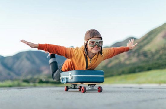 Jeune garçon habillé en aviateur juché sur une valise montée sur une planche à roulettes